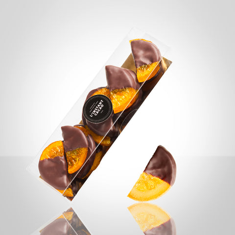 Boite de demi tranches d'oranges confites trempées en partie dans du chocolat noir et demi tranche d'orange trempée en partie dans du chocolat noir posée à côté